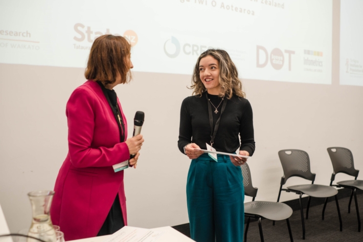 Tori Diamond is awarded the PANZ Newell Prize by PANZ President Rosemary Goodyear. A young woman with sandy wavy hair, black top and a blue skirt smiles as a woman in a red jacket holding a microphone turns towards her.